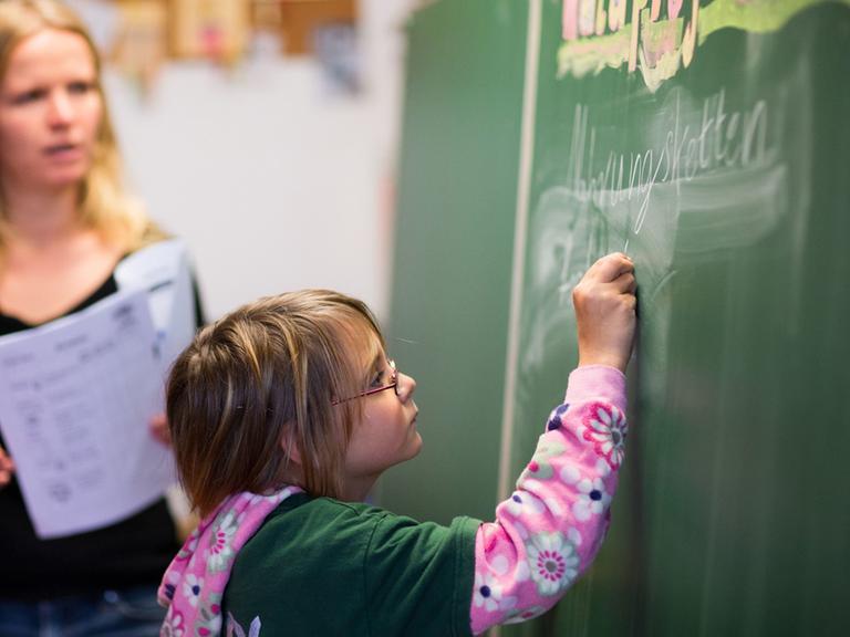 Die neunjährige Cora schreibt, beobachtet von Klassenlehrerin Anne John, in der Freien Schule in Bröbberow an die Schultafel.
