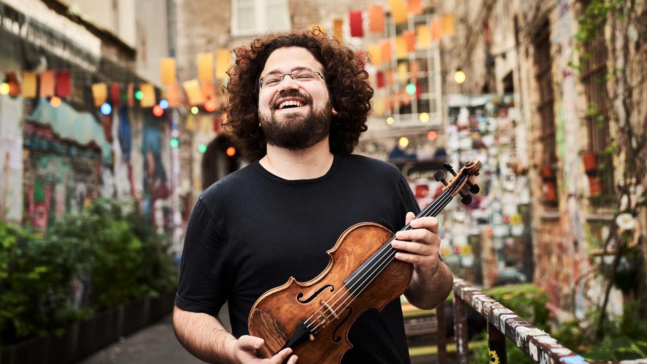 Der Geiger steht inmitten einer bunten Straße, hält seine Geige in beiden Händen und lacht.