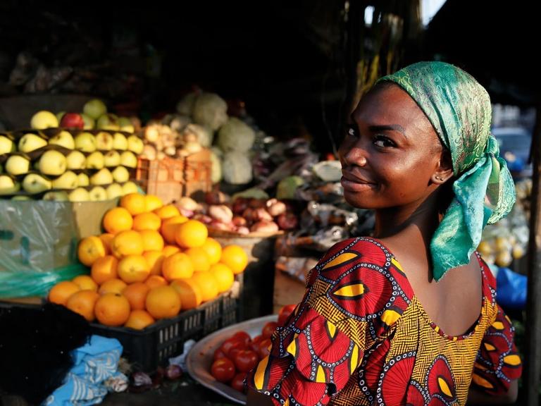 Eine Obstverkäuferin blickt über die Schulter in die Kamera. Im Hintergrund sind Obststände mit reifen Früchten zu sehen.