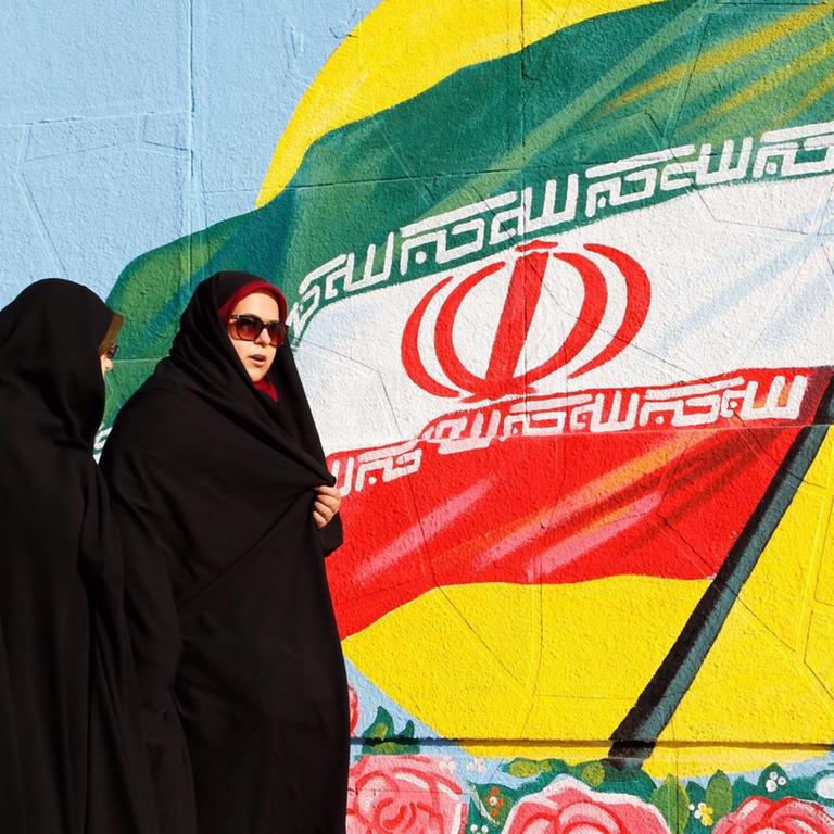 Zwei iranische Frauen in Teheran vor einer iranischen Flagge, die als Graffiti an eine Wand gesprüht ist