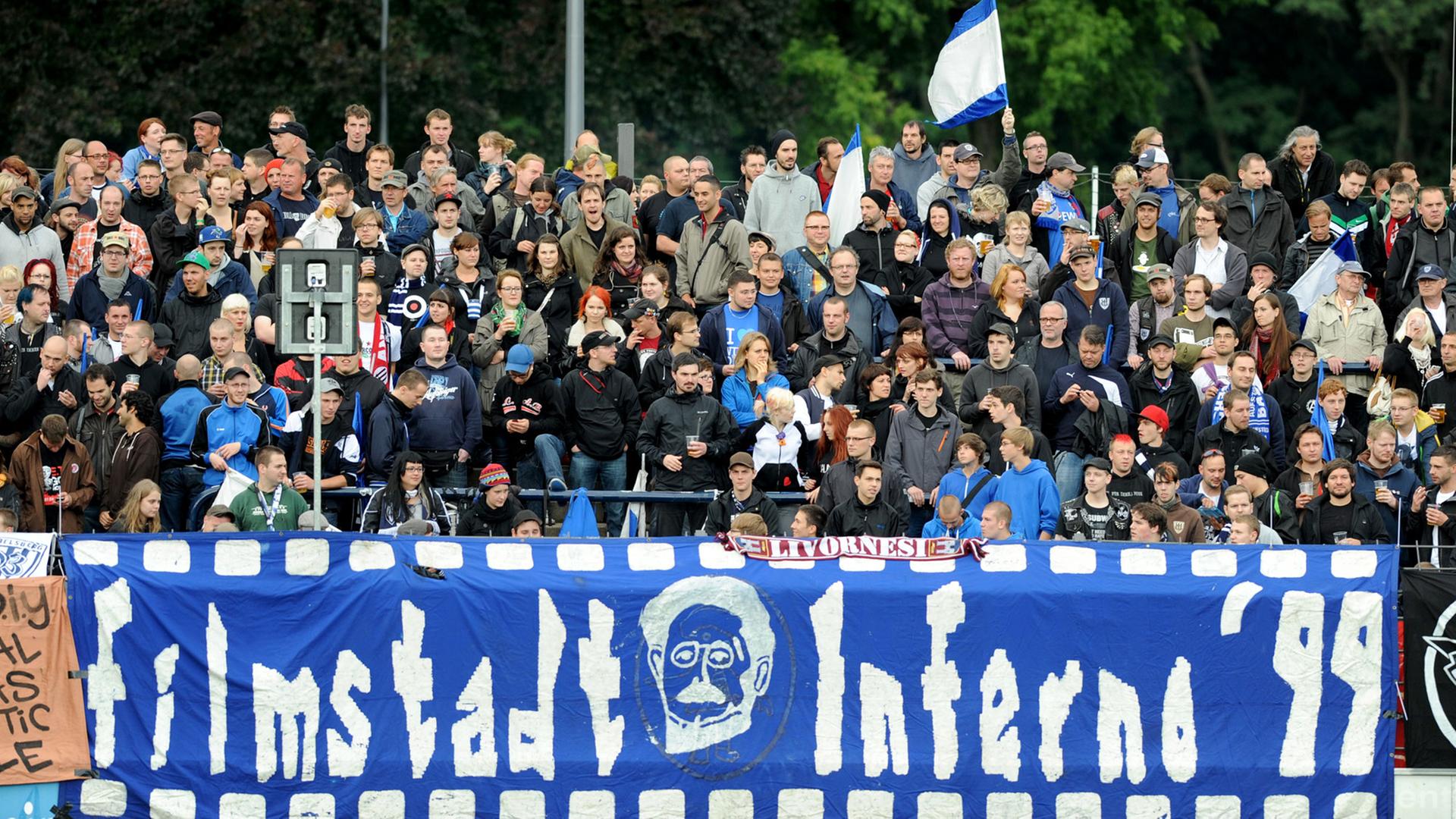 Fans des SV Babelsberg 03 stehen auf einer Stadiontribüne, eine blau weiße Fahne ist zu sehen und im Vordergrund ein Banner mit der Aufschrift "Filmstadt Inferno '99". Fans des SV Babelsberg 03 stehen auf einer Stadiontribüne, eine blau weiße Fahne ist zu sehen und im Vordergrund ein Banner mit der Aufschrift "Filmstadt Inferno '99".