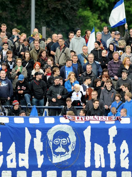 Fans des SV Babelsberg 03 stehen auf einer Stadiontribüne, eine blau weiße Fahne ist zu sehen und im Vordergrund ein Banner mit der Aufschrift "Filmstadt Inferno '99".