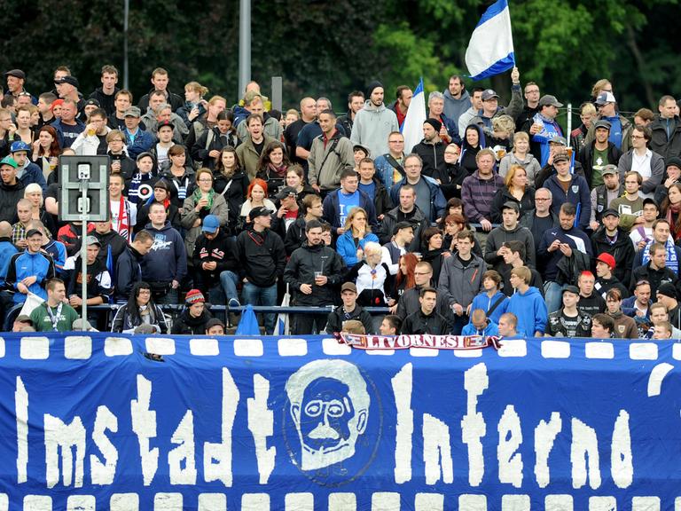Fans des SV Babelsberg 03 stehen auf einer Stadiontribüne, eine blau weiße Fahne ist zu sehen und im Vordergrund ein Banner mit der Aufschrift "Filmstadt Inferno '99".