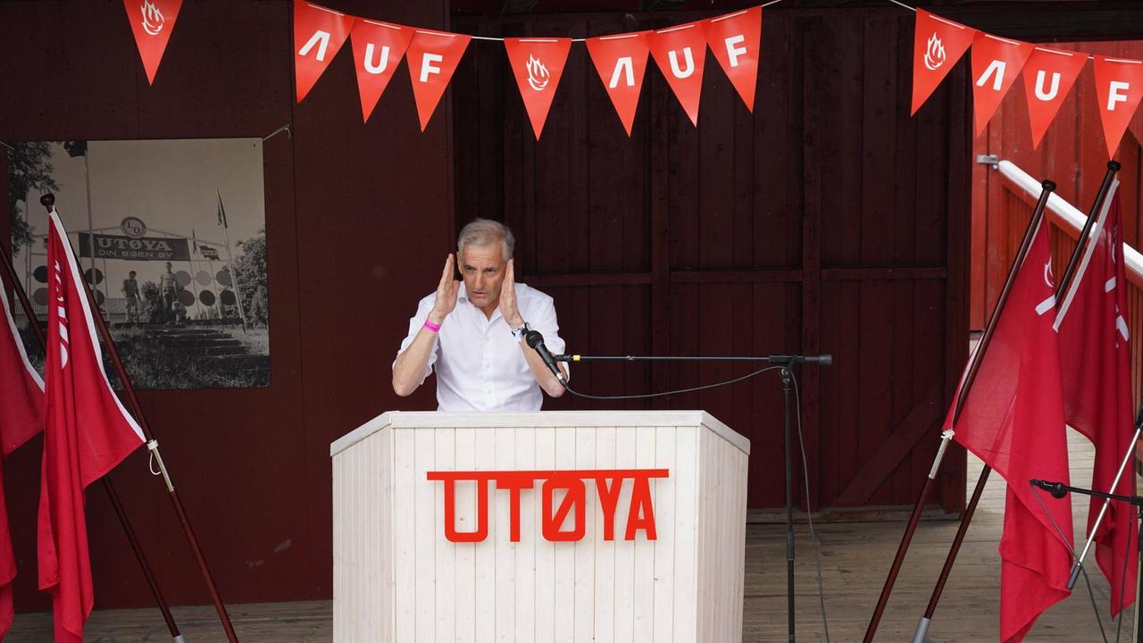 Jonas Gahr Støre steht vor einem weißen, mit Holz verkleidetem Rednerpult, an dem in roter Schrift Utøya steht. Die Bühne schmücken mehrere Wimpelketten, auf denen 'AUF' steht, die sozialdemokratische Arbeiterpartei Norwegens.
