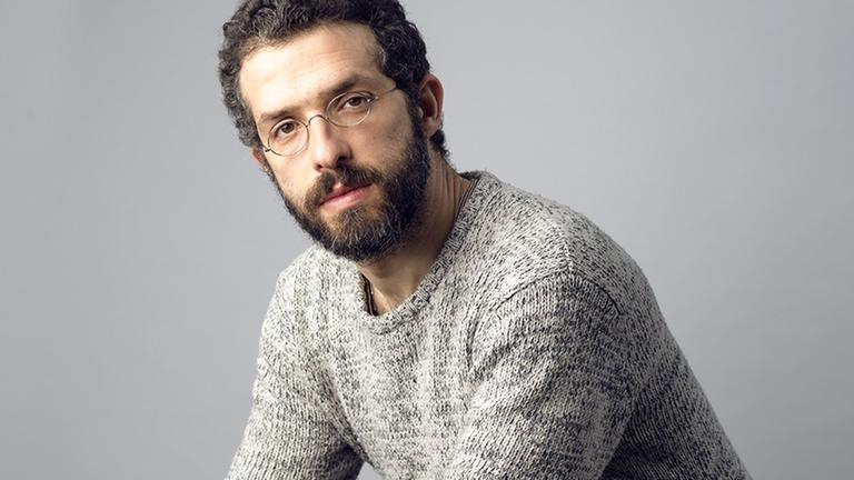 Omer Meir Wellber sitzt auf einem Hocker, trägt einen grauen Strickpullover und blickt in die Kamera