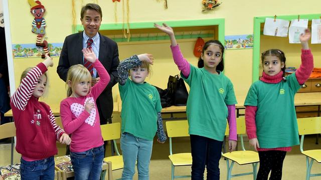 Brandenburgs Bildungsminister Günter Baaske steht hinter fünf Erstklässlern, die winken und tanzen - unter ihnen zwei syrische Mädchen.