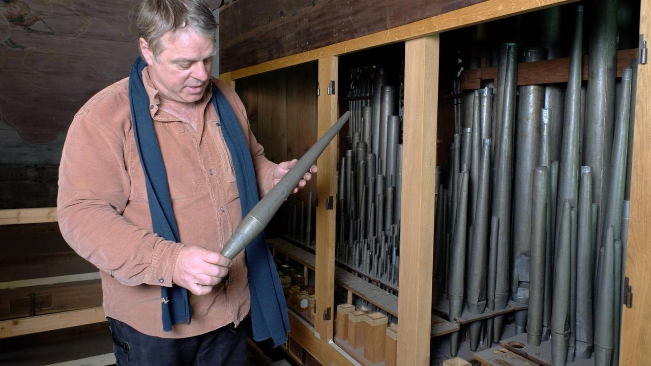 Ein Mann steht neben einem offenen Orgelkasten, in dem verschieden große, verstaubte Orgelpfeifen stehen. Er hat eine der alten Zinnpfeifen in der Hand und betrachtet ihren Zustand. Ein Mann steht neben einem offenen Orgelkasten, in dem verschieden große, verstaubte Orgelpfeifen stehen. Er hat eine der alten Zinnpfeifen in der Hand und betrachtet ihren Zustand.