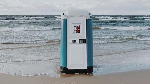 Ein transportables Toilettenhäuschen steht in der Meeresbrandung am Strand.
