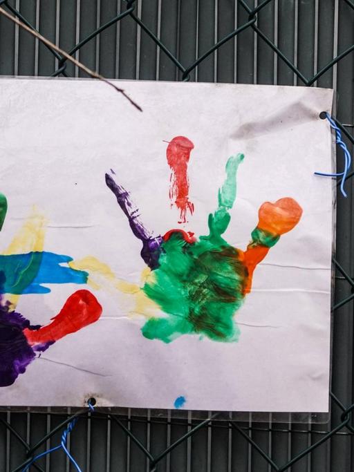 An einem Zaun hängt ein Blatt Papier mit dem vielfarbigen Abdruck zweier Kinderhände.