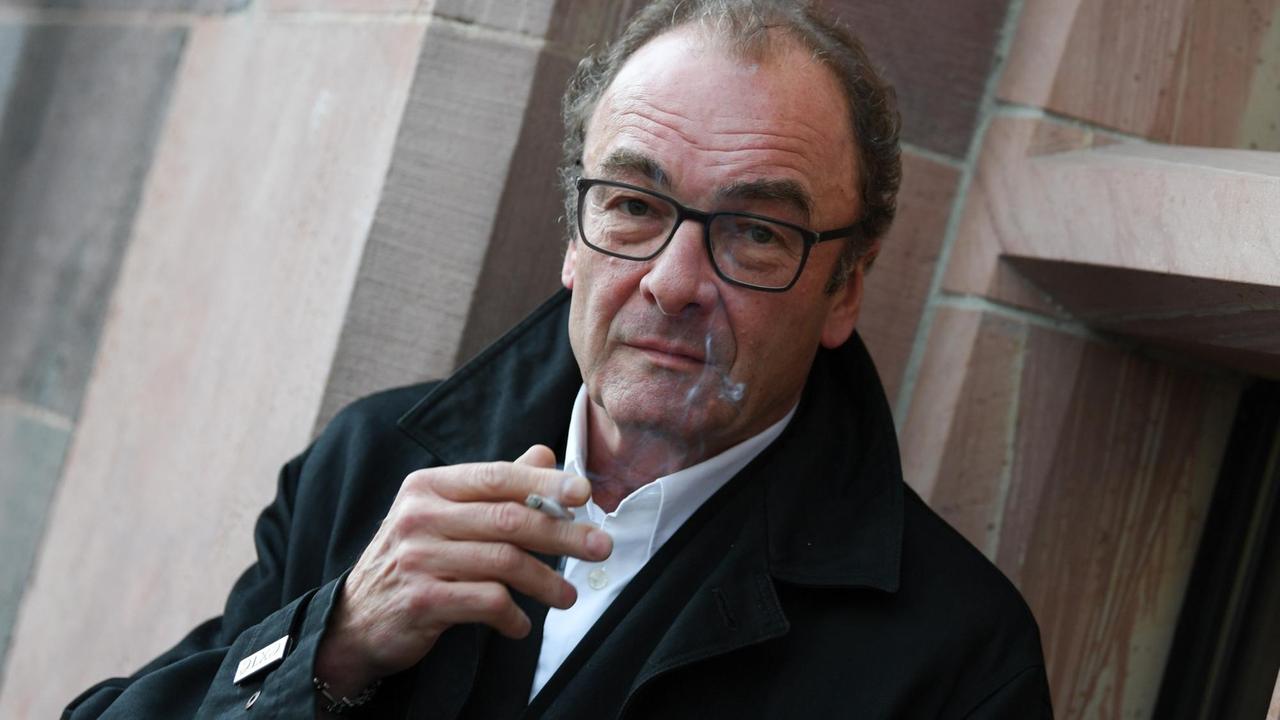 Robert Menasse hält eine Zigarette in der Hand und schaut in die Kamera. Robert Menasse hält eine Zigarette in der Hand und schaut in die Kamera.