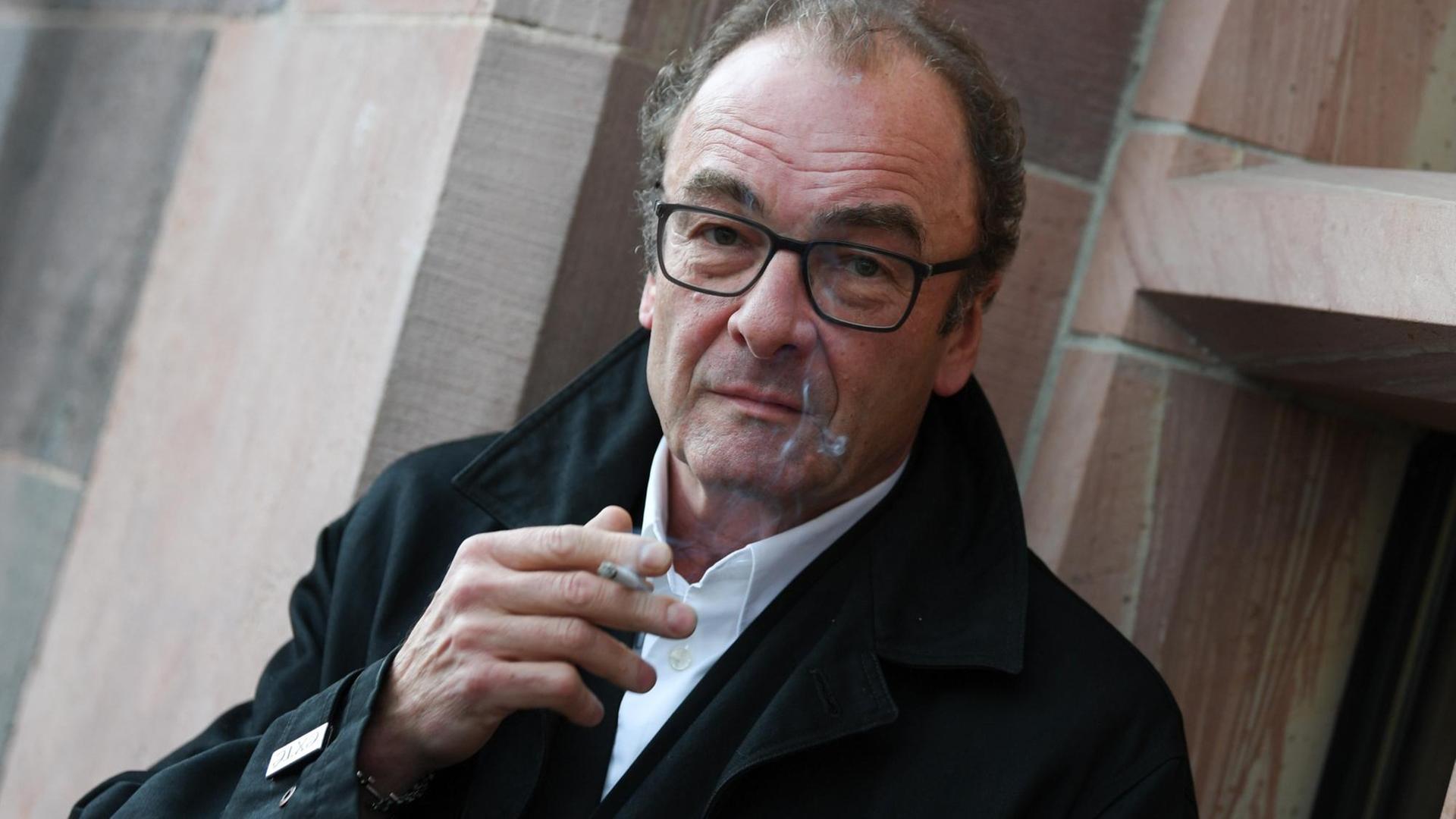Robert Menasse hält eine Zigarette in der Hand und schaut in die Kamera. Robert Menasse hält eine Zigarette in der Hand und schaut in die Kamera.