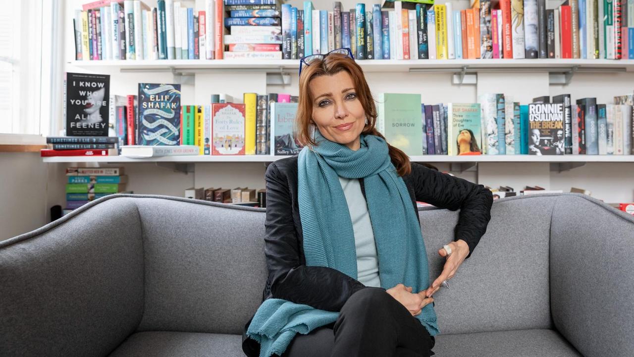 Die Schriftstellerin Elif Shafak sitzt auf einem grauen Sofa. Im Hintergrund ist ein Bücherregal zu sehen. Die Schriftstellerin Elif Shafak sitzt auf einem grauen Sofa. Im Hintergrund ist ein Bücherregal zu sehen.