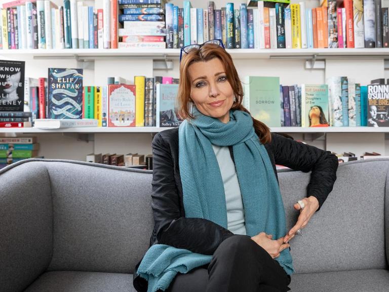 Die Schriftstellerin Elif Shafak sitzt auf einem grauen Sofa. Im Hintergrund ist ein Bücherregal zu sehen.