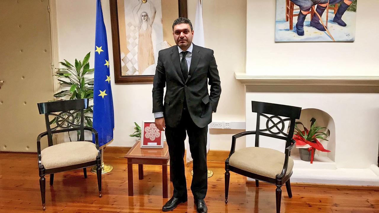 Zyperns Innenminister Konstantinos Petrides steht im dunklen Anzug in seinem Amtszimmer. Im Hintergrund Bilder an der Wand sowie ein zyprische und eine EU-Flagge.