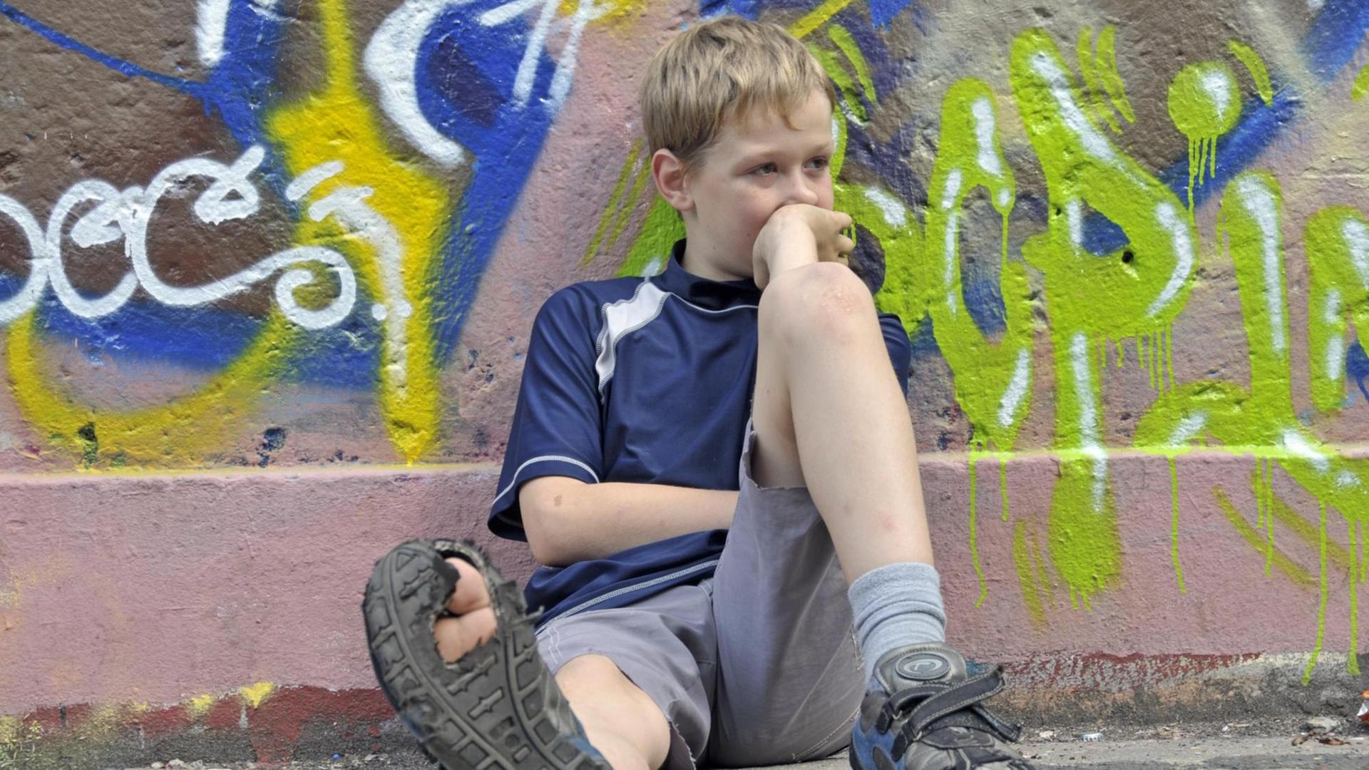 Lustloser neunjähriger Junge sitzt vor einer Graffiti-Wand, einen Fuß von sich gestreckt, sodass die kaputte Sohle des Schuhs sichtbar wird. (Symbolbild)
