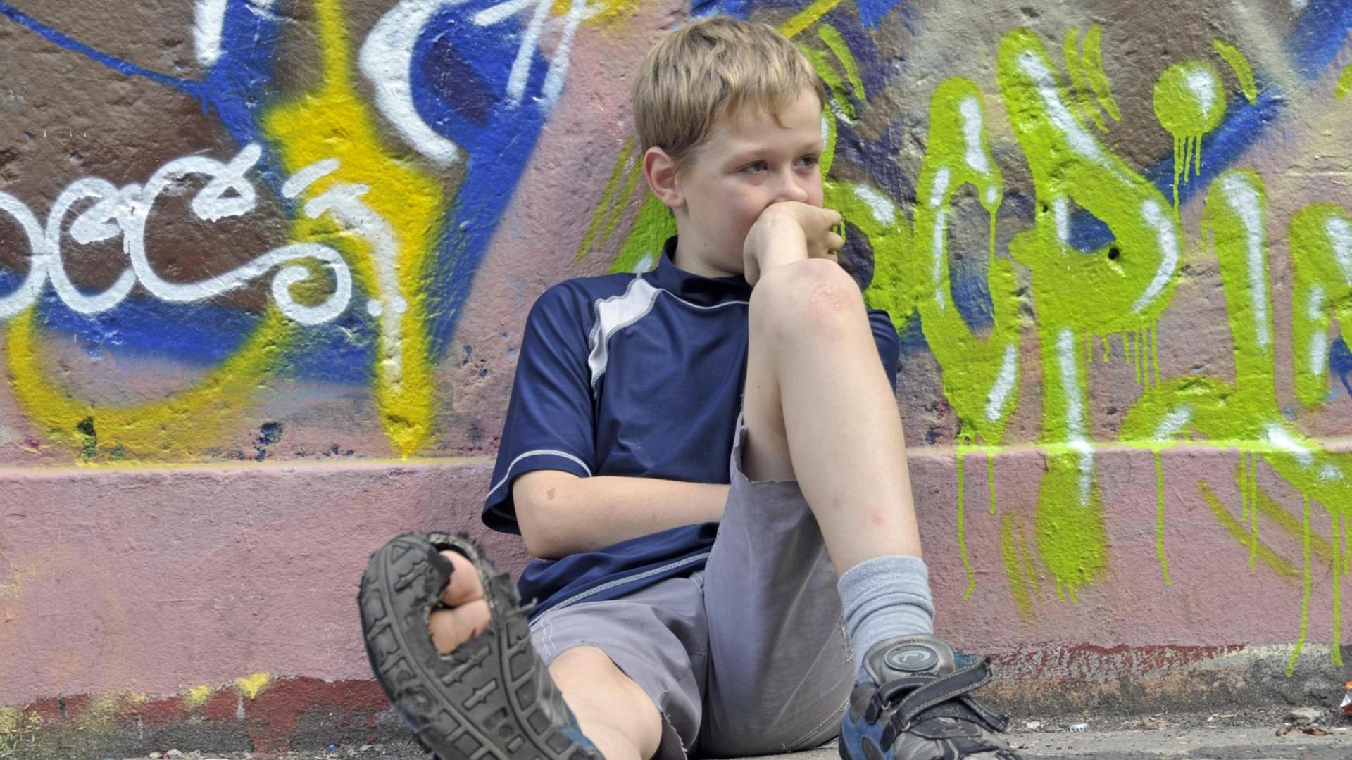 Lustloser neunjähriger Junge sitzt vor einer Graffiti-Wand, einen Fuß von sich gestreckt, sodass die kaputte Sohle des Schuhs sichtbar wird. (Symbolbild) Lustloser neunjähriger Junge sitzt vor einer Graffiti-Wand, einen Fuß von sich gestreckt, sodass die kaputte Sohle des Schuhs sichtbar wird. (Symbolbild)