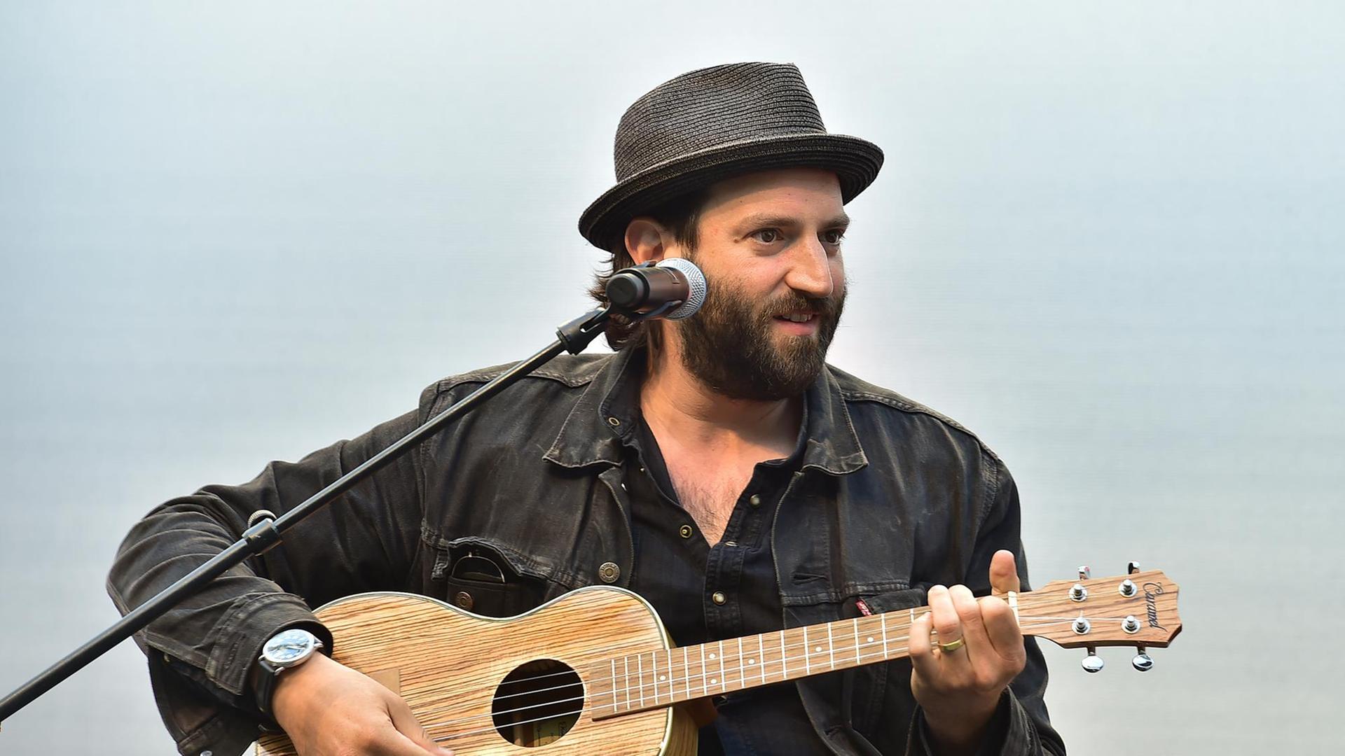 Der Musiker Daniel Kahn sitzt mit schwarzem Hemd, schwarzem Hut und einer Ukulele hinter einem Mikrofon.
