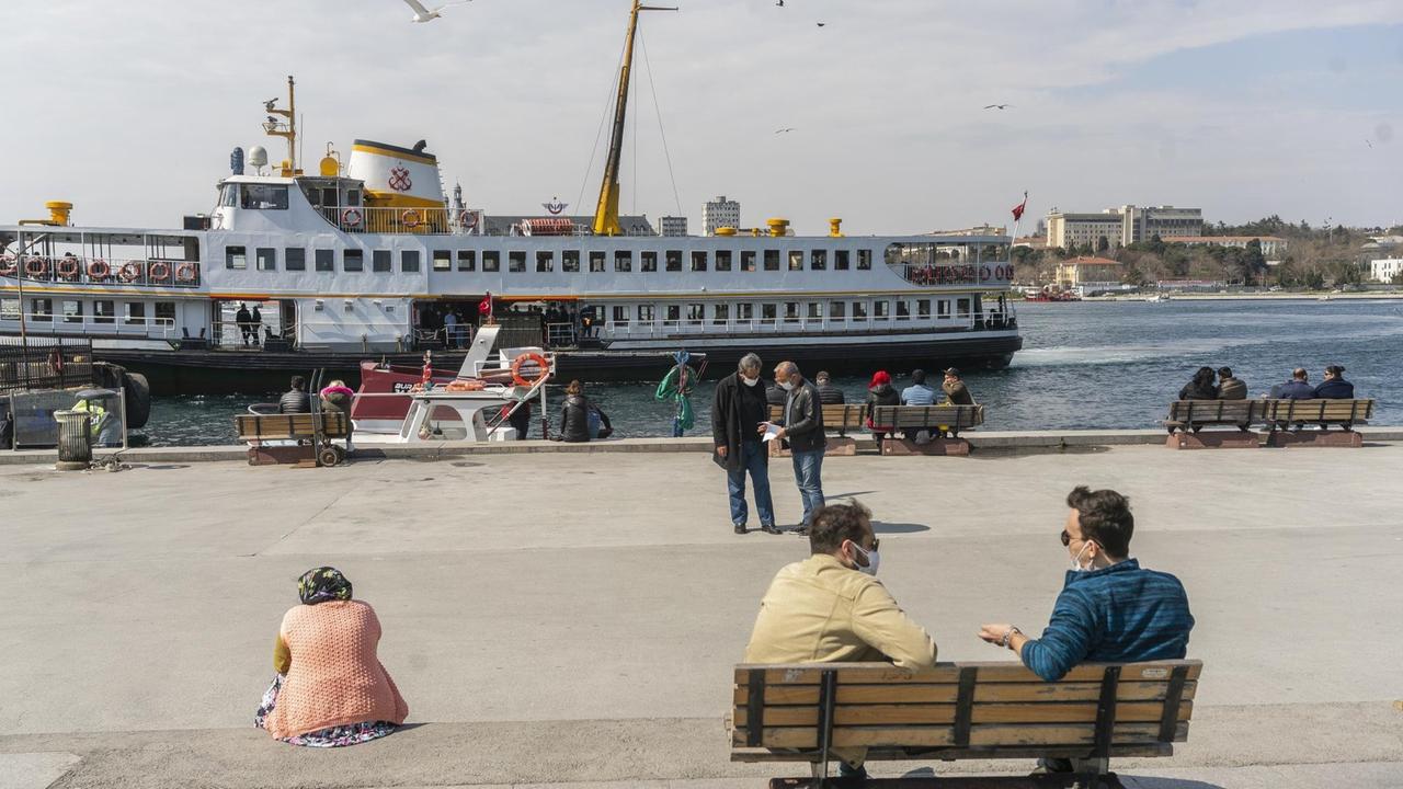 Menschen sitzen am Ufer auf Bänken, im Hintergrund ein Ausflugsdampfer. Menschen sitzen am Ufer auf Bänken, im Hintergrund ein Ausflugsdampfer.
