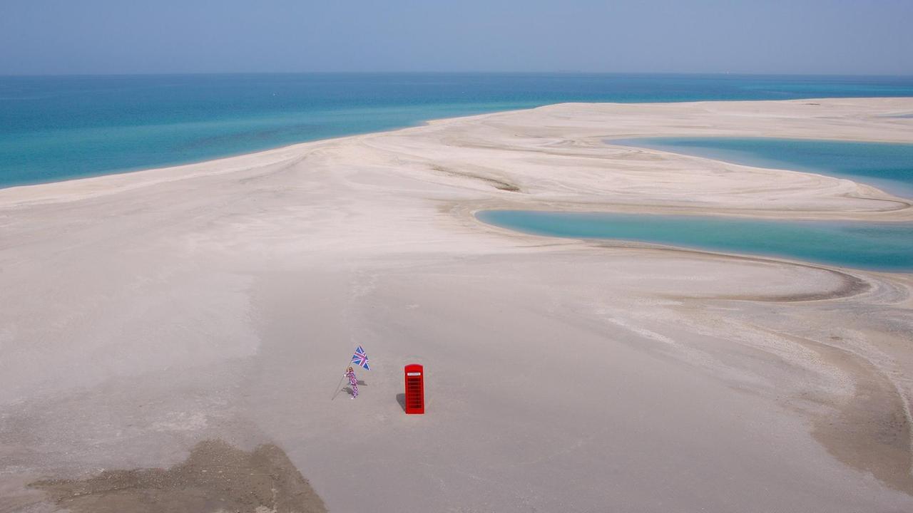 In Vogelperspektive ist eine flache Sandfläche zu erkennen umgeben von türkisfarbenem Wasser. Im Vordergrund steht eine rote, typisch britische Telefonzelle, links daneben ein Mann in Union-Jack Anzug und Flagge. In Vogelperspektive ist eine flache Sandfläche zu erkennen umgeben von türkisfarbenem Wasser. Im Vordergrund steht eine rote, typisch britische Telefonzelle, links daneben ein Mann in Union-Jack Anzug und Flagge.