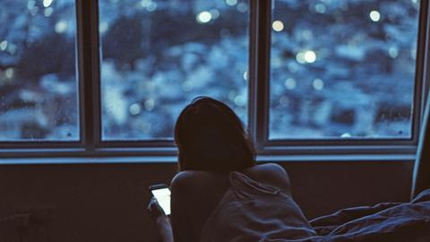 Eine junge Frau liegt bäuchlings im Bett und schaut auf ihr Smartphone.