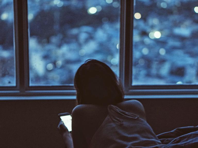 Eine junge Frau liegt bäuchlings im Bett und schaut auf ihr Smartphone.