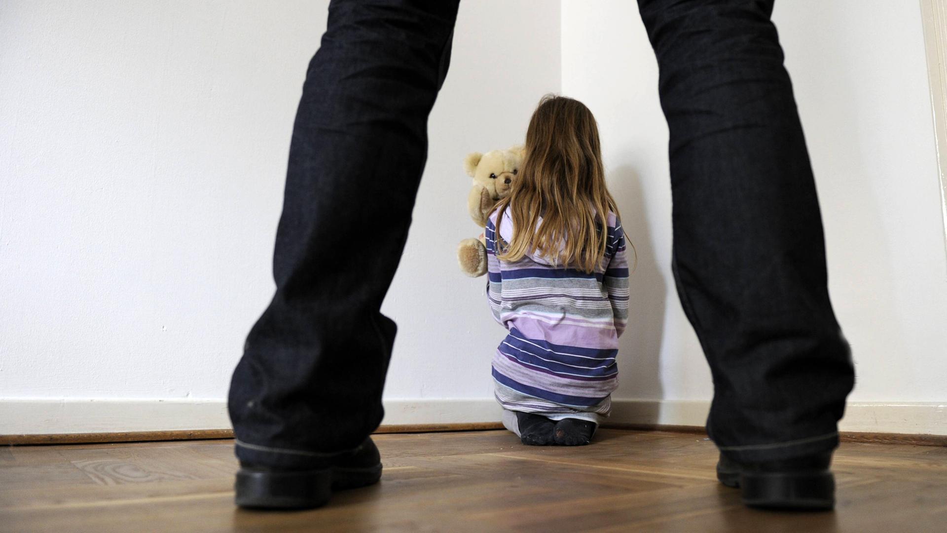 Symbolfoto zum Thema Kindesmissbrauch: Man sieht ein Mädchen in einer Zimmerecke mit Teddy von hinten und im Vordergrund die Beine eines Mannes Symbolfoto zum Thema Kindesmissbrauch: Man sieht ein Mädchen in einer Zimmerecke mit Teddy von hinten und im Vordergrund die Beine eines Mannes