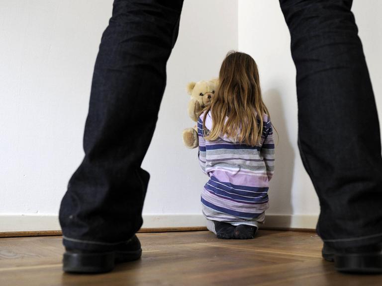 Symbolfoto zum Thema Kindesmissbrauch: Man sieht ein Mädchen in einer Zimmerecke mit Teddy von hinten und im Vordergrund die Beine eines Mannes