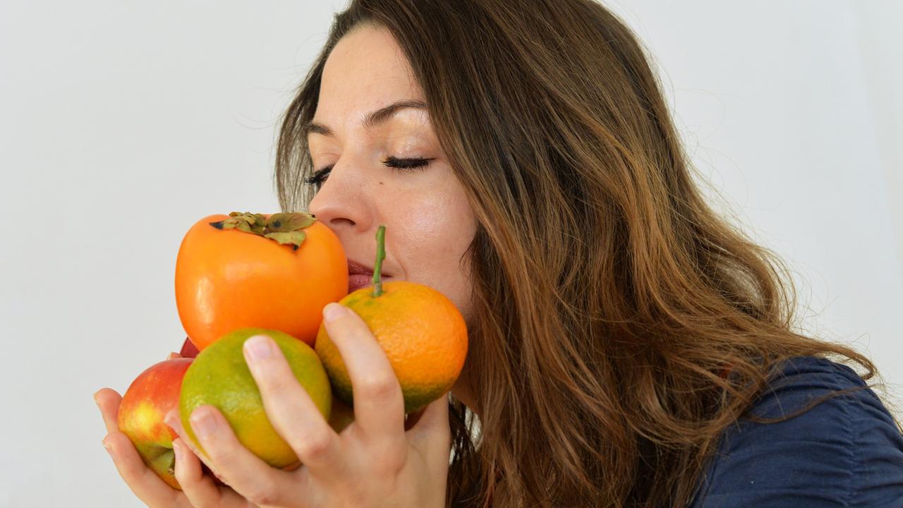 Eine junge Frau hält Äpfel, Mandarinen und eine Kaki in den Händen und riecht daran.