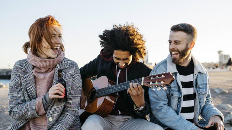 Three happy friends with guitar sitting outdoors at sunset model released Symbolfoto PUBLICATIONxINxGERxSUIxAUTxHUNxONLY JRFF02654