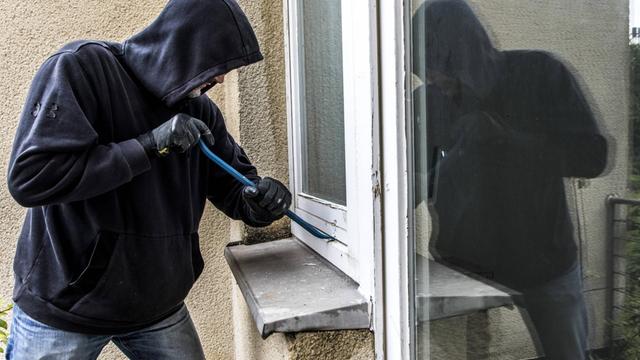 Ein Mann mit Hoodie bricht mit Werkzeug einen Fensterrahmen auf