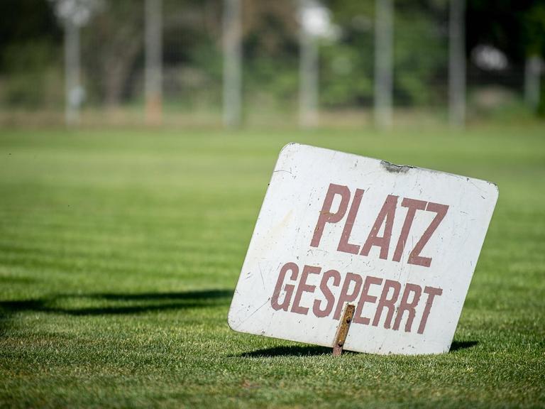 Ein Schild mit der Aufschrift "Platz gesperrt" steckt im Rasen eines Vereinsspielfeldes.
