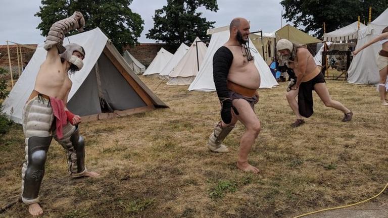 Drei Männer stehen auf einer Wiese und ertüchtigen sich körperlich. Im Hintergrund sind Zelte aufgestellt. Drei Männer stehen auf einer Wiese und ertüchtigen sich körperlich. Im Hintergrund sind Zelte aufgestellt.
