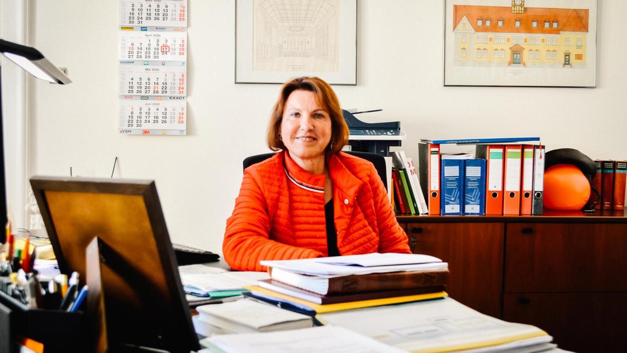 Ines Hübner (SPD), Bürgermeisterin von Velten, sitzt an einem Schreibtisch. Im Vordergrund die Rückansicht eines Bilderrahmens, im Hintergrund Aktenordner und ein Bild des Rathauses der Stadt. Ines Hübner (SPD), Bürgermeisterin von Velten, sitzt an einem Schreibtisch. Im Vordergrund die Rückansicht eines Bilderrahmens, im Hintergrund Aktenordner und ein Bild des Rathauses der Stadt.