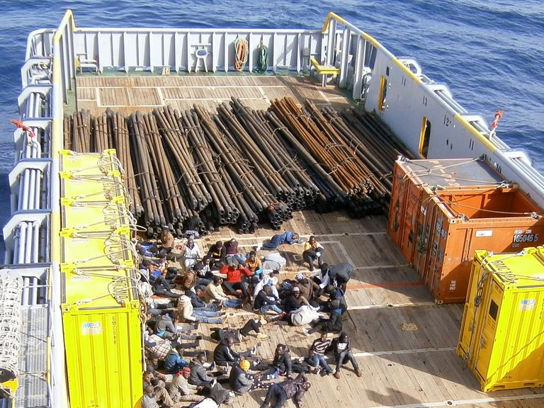 Im hinteren Teil eines Frachtschiffes befinden sich Dutzende Flüchtlinge, hinter ihnen Stahlträger und einige kleinere Container.