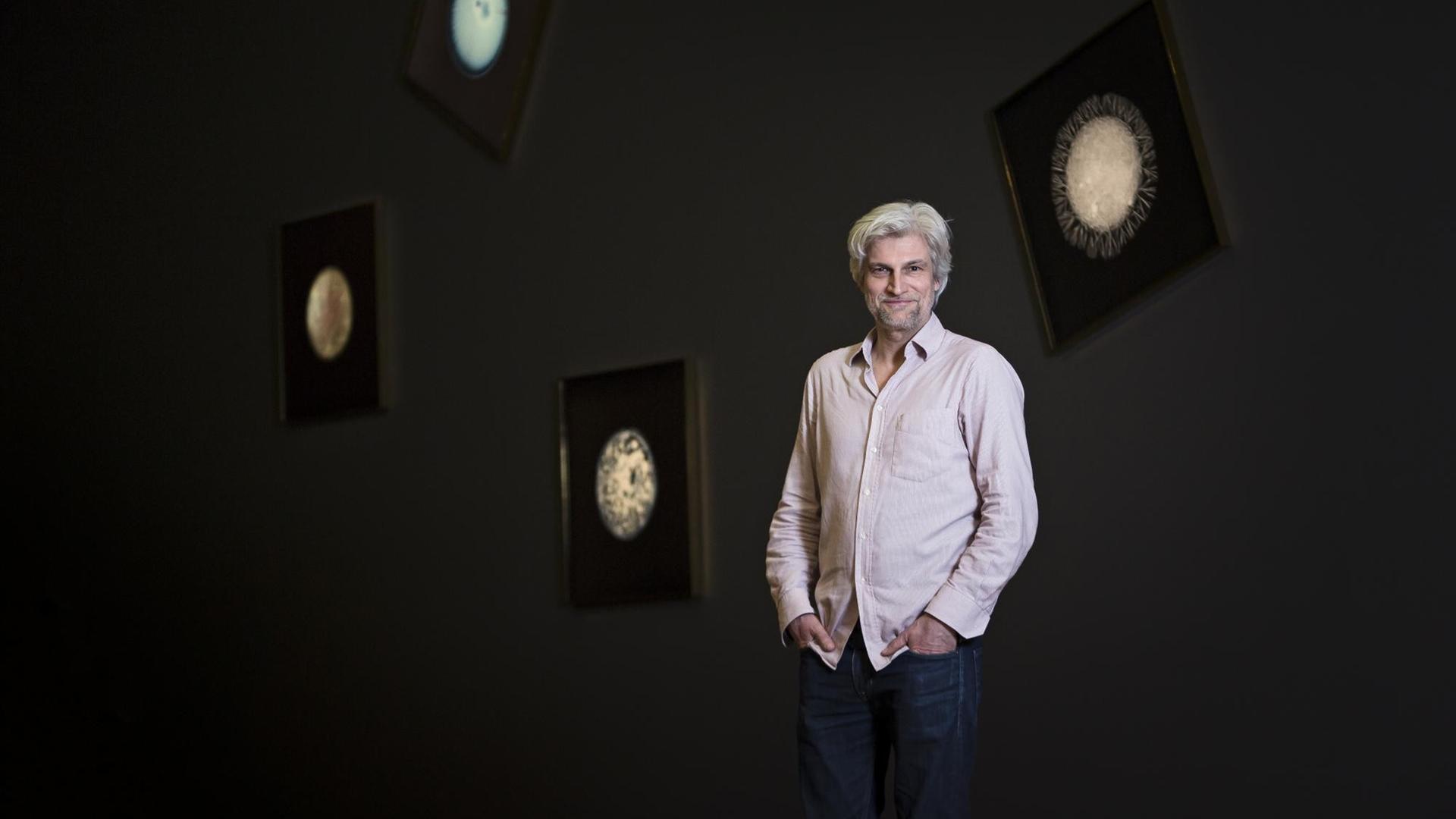 Jörg Völlnagel steht in einer Ausstellung vor einer schwarzen Wand, im Hintergrund hängen Bilder, die im Dunklen zu leuchten scheinen.