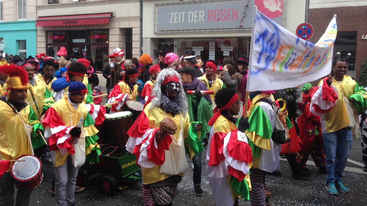 Eine grün-gelb gekleidete Karnevalsgruppe läuft auf einer Straße in einem Umzug mit, mit einem Transparent, auf dem steht: "Mer losse uns Flüchtlinge in Kölle". Eine grün-gelb gekleidete Karnevalsgruppe läuft auf einer Straße in einem Umzug mit, mit einem Transparent, auf dem steht: "Mer losse uns Flüchtlinge in Kölle".