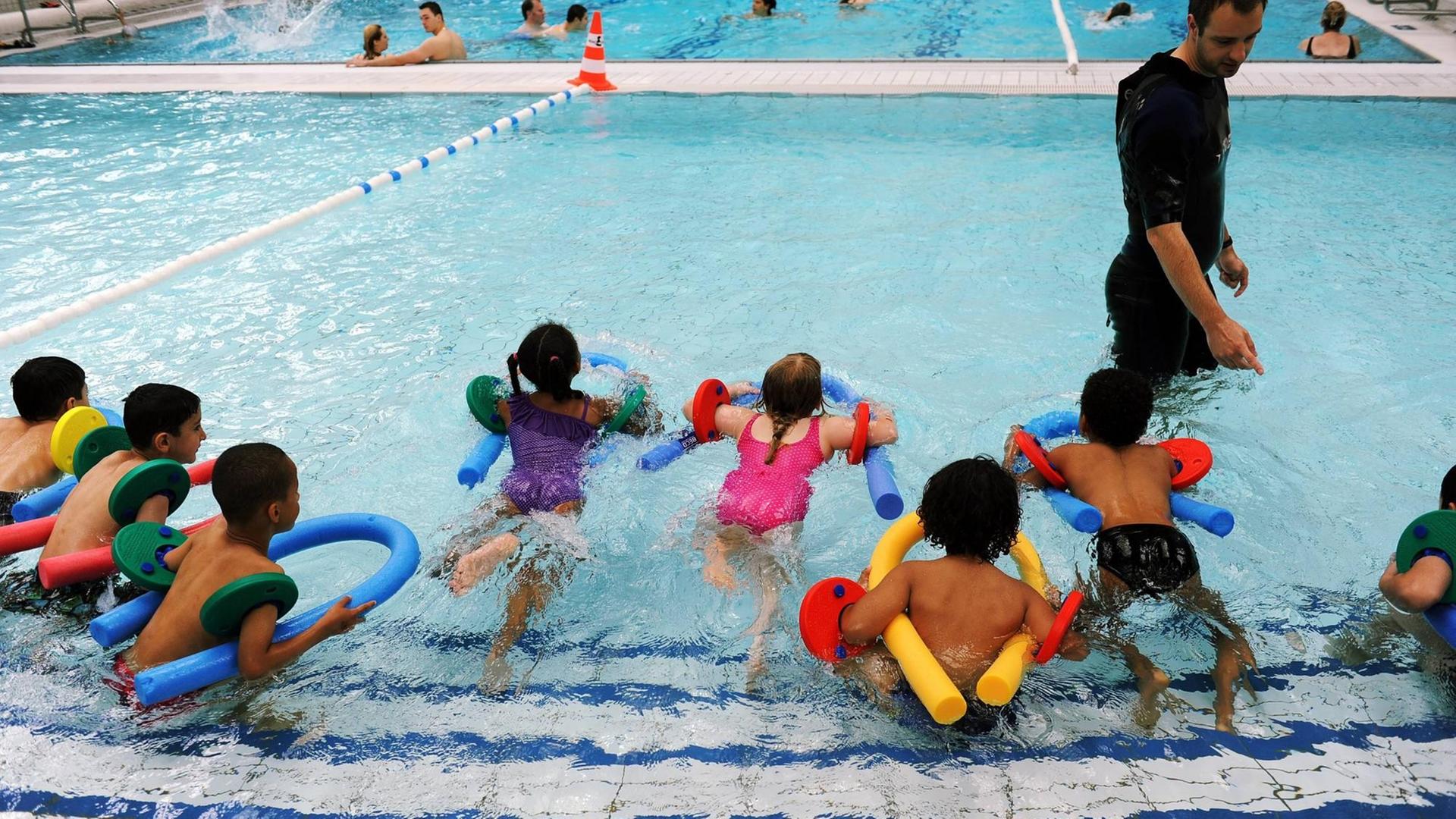 In einem Schwimmbecken stoßen sich knapp zehn Kinder vom Beckenrand im Nichtschwimmerbereich ab. Sie haben Schwimmnudeln. Ein Schwimmlehrer steht im Wasser in schwarzem Short und Shirt vor ihnen. In einem Schwimmbecken stoßen sich knapp zehn Kinder vom Beckenrand im Nichtschwimmerbereich ab. Sie haben Schwimmnudeln. Ein Schwimmlehrer steht im Wasser in schwarzem Short und Shirt vor ihnen.
