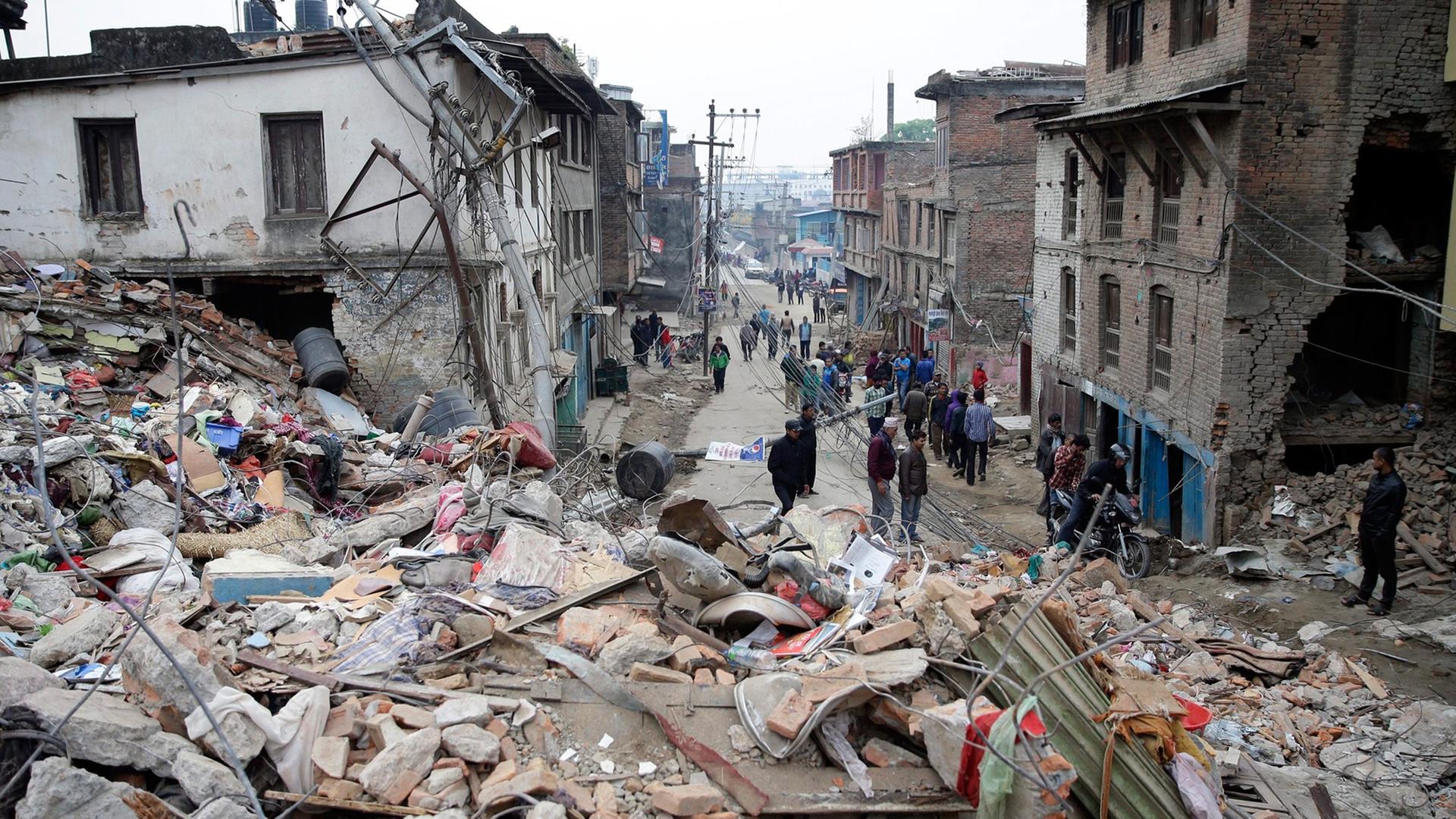 Trümmer eines eingestürzten Hauses in Kathmandu, im Hintergrund der Blick in eine Straße mit zerstörten Häusern und einigen Menschen. Trümmer eines eingestürzten Hauses in Kathmandu, im Hintergrund der Blick in eine Straße mit zerstörten Häusern und einigen Menschen.