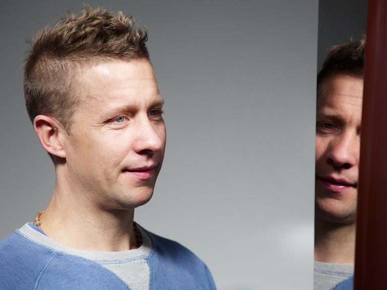 Portrait von Jeppe Hein, der vor einem Spiegel steht und sich teilweise darin spiegelt.