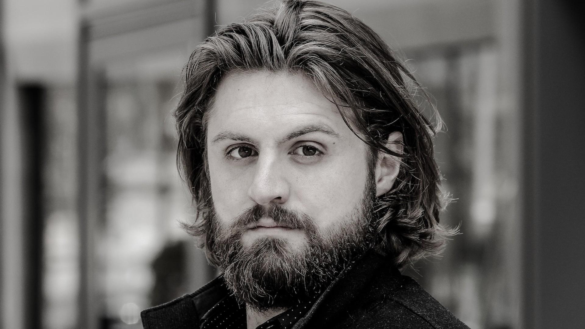 Eine junger Mann mit Bart und längeren Haaren blickt ernst in die Kamera. Es ist der Bariton Konstantin Krimmel. Eine junger Mann mit Bart und längeren Haaren blickt ernst in die Kamera. Es ist der Bariton Konstantin Krimmel.