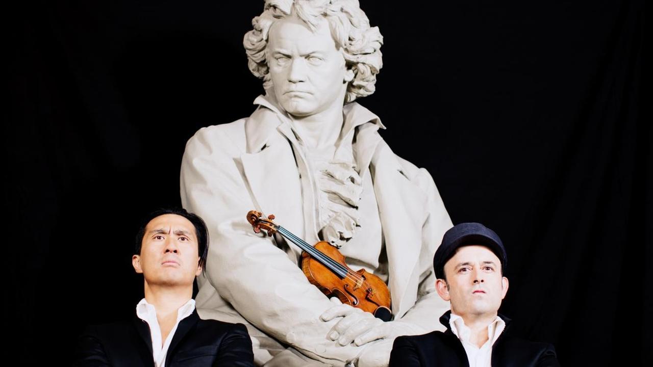 Zwei Männer stehen vor einer Beethovenstatue mit verschränkten Armen und ernsten Blicken. Beethoven hält eine Geige im Arm. Die beiden Männer sind das Comedy-Duo Igudesman und Joo. Zwei Männer stehen vor einer Beethovenstatue mit verschränkten Armen und ernsten Blicken. Beethoven hält eine Geige im Arm. Die beiden Männer sind das Comedy-Duo Igudesman und Joo.