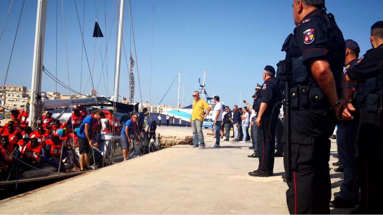 Zu sehen ist ein Schiff mit Migranten an  Bord, die rote Rettungswesten tragen. Das Schiff liegt an einem Dock im Hafen. Daneben stehen Polizisten an Land.