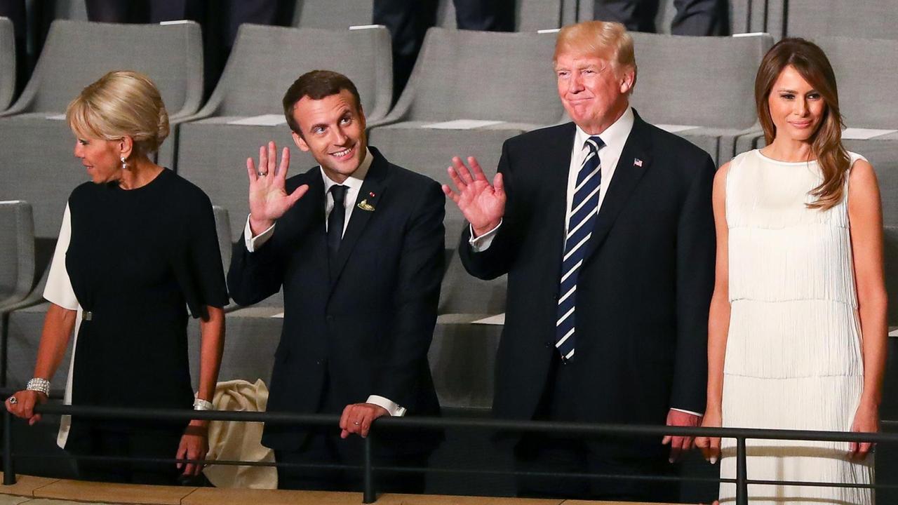 Die vier stehen nebeneinander vor ihren Plätzen an einem Geländer: v. l.n.r. Frau und Herr Macron, Herr und Frau Trump. Die beiden Präsidenten winken lächelnd. Die vier stehen nebeneinander vor ihren Plätzen an einem Geländer: v. l.n.r. Frau und Herr Macron, Herr und Frau Trump. Die beiden Präsidenten winken lächelnd.