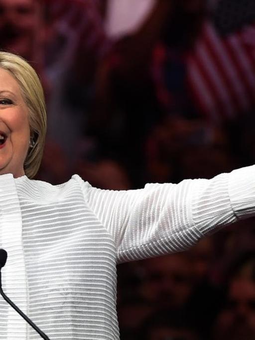 Hillary Clinton steht vor jubelnden Anhängern und breitet lächelnd die Arme aus.
