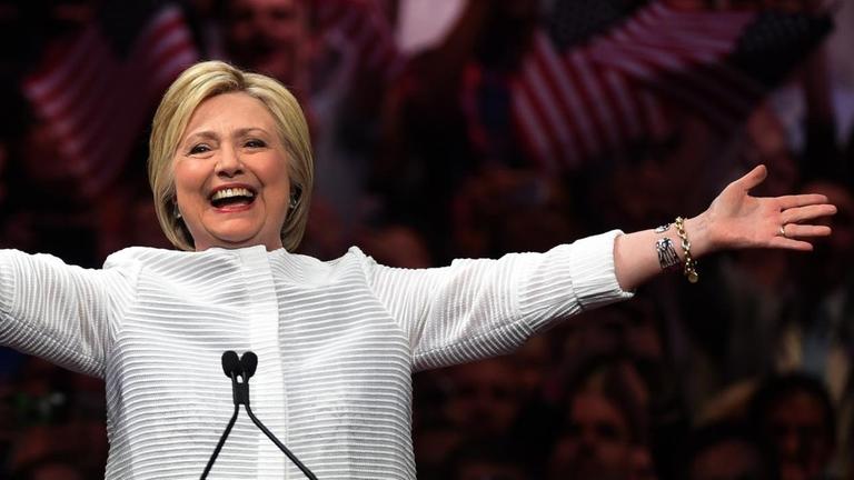 Hillary Clinton steht vor jubelnden Anhängern und breitet lächelnd die Arme aus.