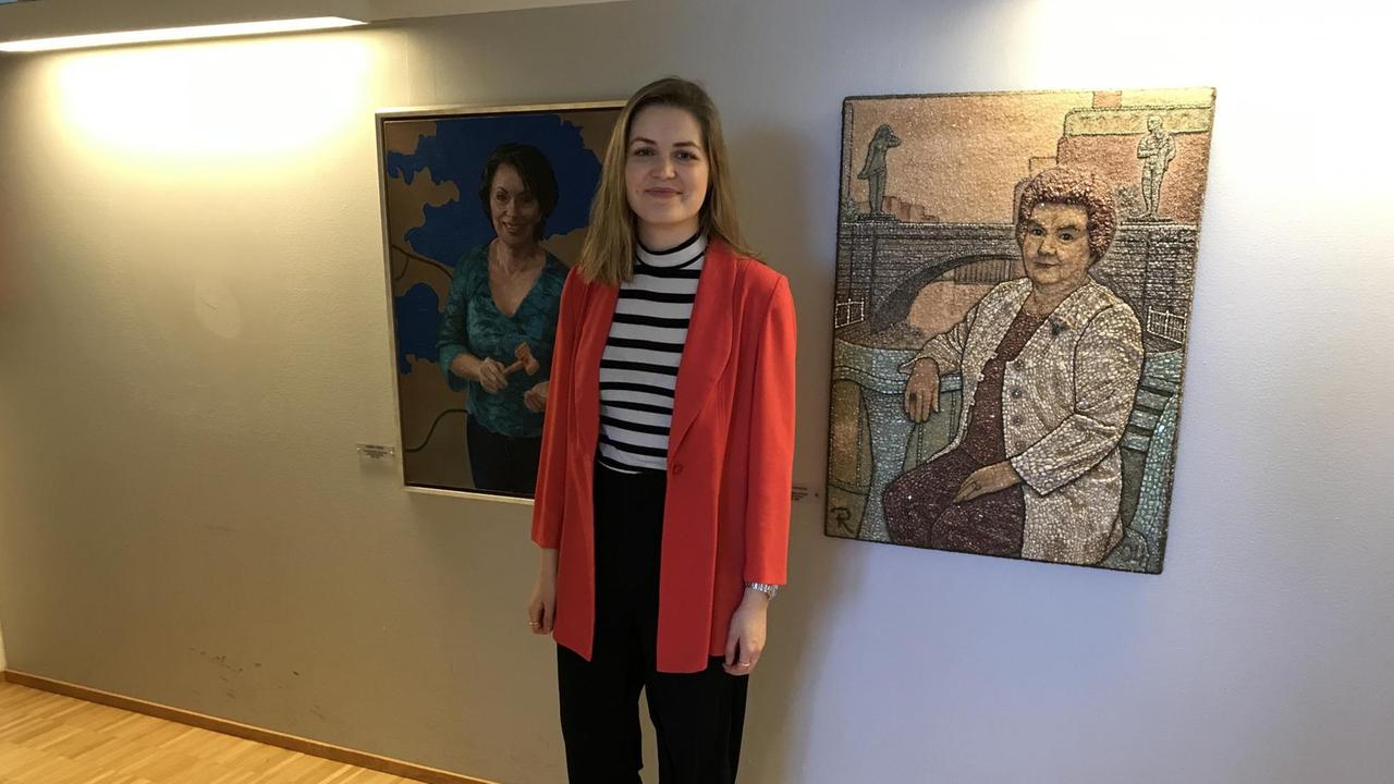 Suvi Mäkelainen, Parlamentskandidatin der Zentrumspartei in Tampere, steht in einer roten langen Strickjacke und schwarzer Hose vor zwei Porträts von Frauen. Suvi Mäkelainen, Parlamentskandidatin der Zentrumspartei in Tampere, steht in einer roten langen Strickjacke und schwarzer Hose vor zwei Porträts von Frauen.