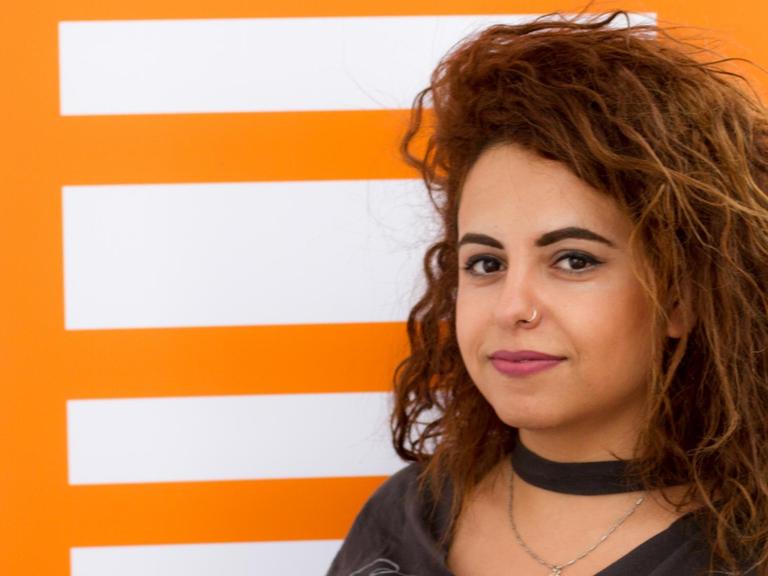 Die syrische Journalistin Hiba Obaid hat lange schwarze Haare und steht vor einer Wand mit dem Deutschlandfunk Kultur Logo.