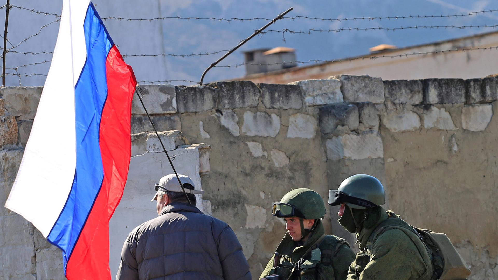Zwei Soldaten in Tarnkleidung ohne Hoheitsabzeichen stehen bei einem Mann mit einer russischen Flagge. Zwei Soldaten in Tarnkleidung ohne Hoheitsabzeichen stehen bei einem Mann mit einer russischen Flagge.