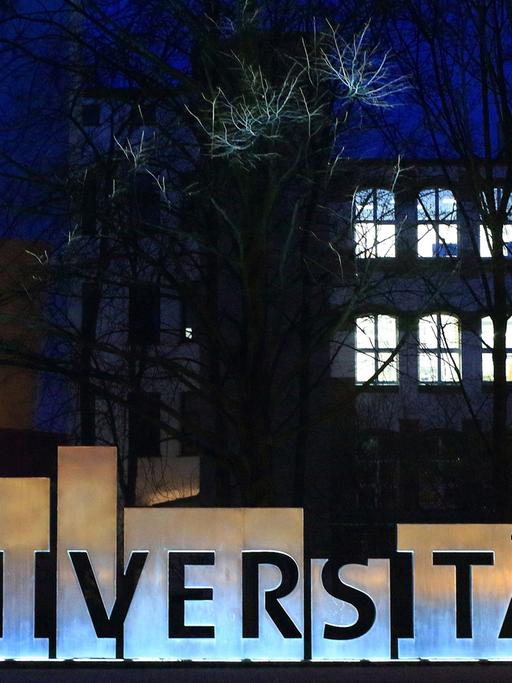 Die Fassaden eines Universitätsgebäudes bei Nacht mit hell erleuchteten Fenstern, im Vordergrund der Schriftzug "Universität".