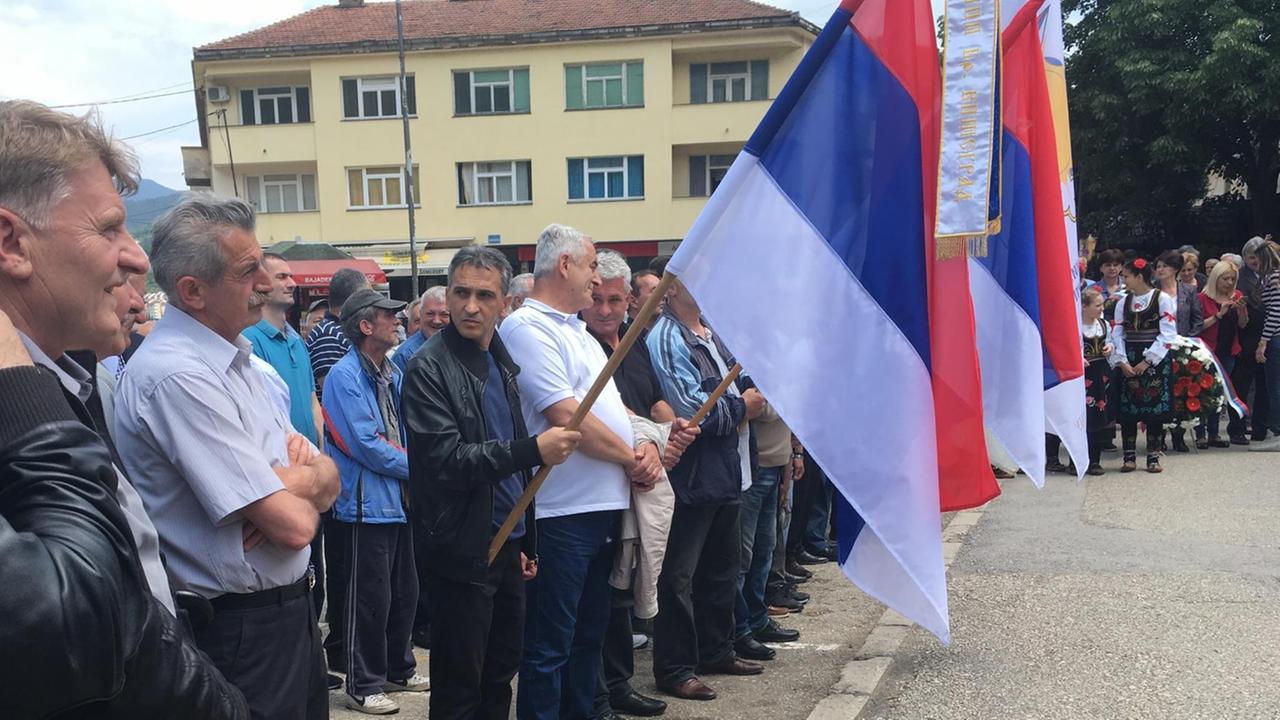 Eine Gruppe von Männern steht vor Vor dem Denkmal für die "Verteidiger der Republika Srpska" in der Nähe der Brücke. Drei der Männer schwenken Fahnen. Eine Gruppe von Männern steht vor Vor dem Denkmal für die "Verteidiger der Republika Srpska" in der Nähe der Brücke. Drei der Männer schwenken Fahnen.